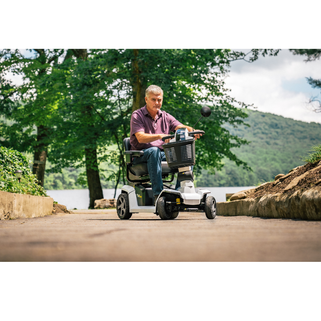A man rides a Pride Mobility Zero Turn 10 HD Scooter along an outdoor path, surrounded by trees, greenery, and a lake in the background on a sunny day.
