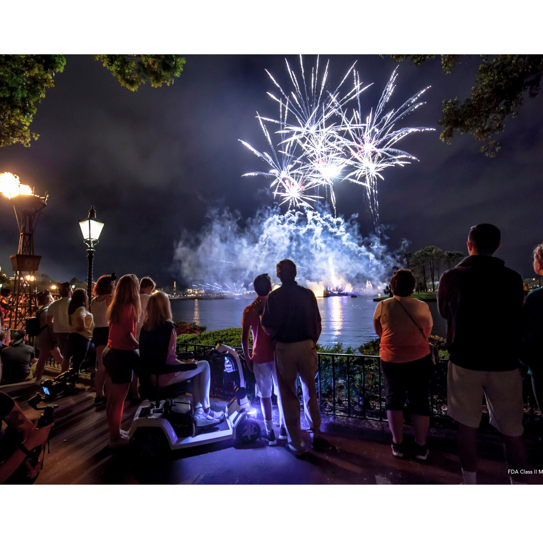 A group of people, some standing and one seated on a Pride Mobility Zero Turn 10 HD Scooter with EZ Turn Technology, watch fireworks light up the night sky over a lake, surrounded by trees and torches.