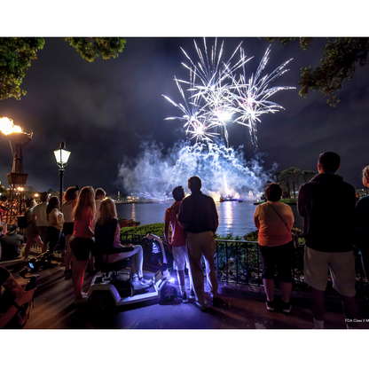 A group of people, some standing and one seated on a Pride Mobility Zero Turn 10 HD Scooter with EZ Turn Technology, watch fireworks light up the night sky over a lake, surrounded by trees and torches.