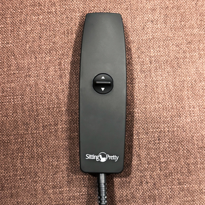 A black remote with up/down arrow buttons, labeled Sitting Pretty, rests on a textured brown surface—it belongs to the Pride Mobility Genesis LC150 Lift Chair Recliner; see product specs for more details.