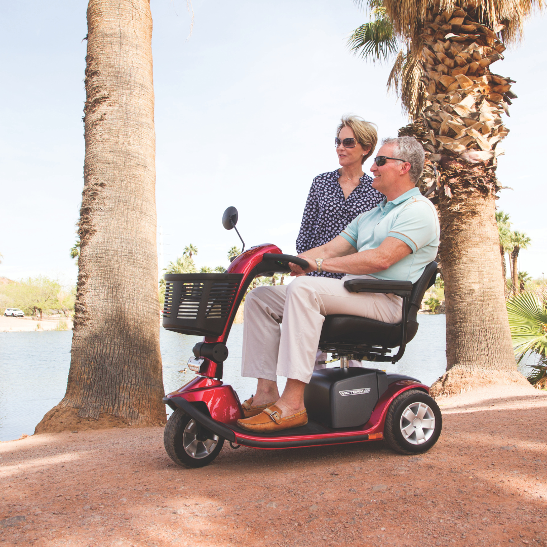 A man sits on a Pride Mobility Victory® 10 Bariatric 3-Wheel Scooter near palm trees and a lake, while a woman stands beside him smiling on a sunny day.