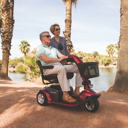 A man rides a red Pride Mobility Victory® 10 Bariatric 3-Wheel Scooter along a sunny path lined with palm trees, while a woman stands behind him. Both are smiling and enjoying the outdoors near a pond.