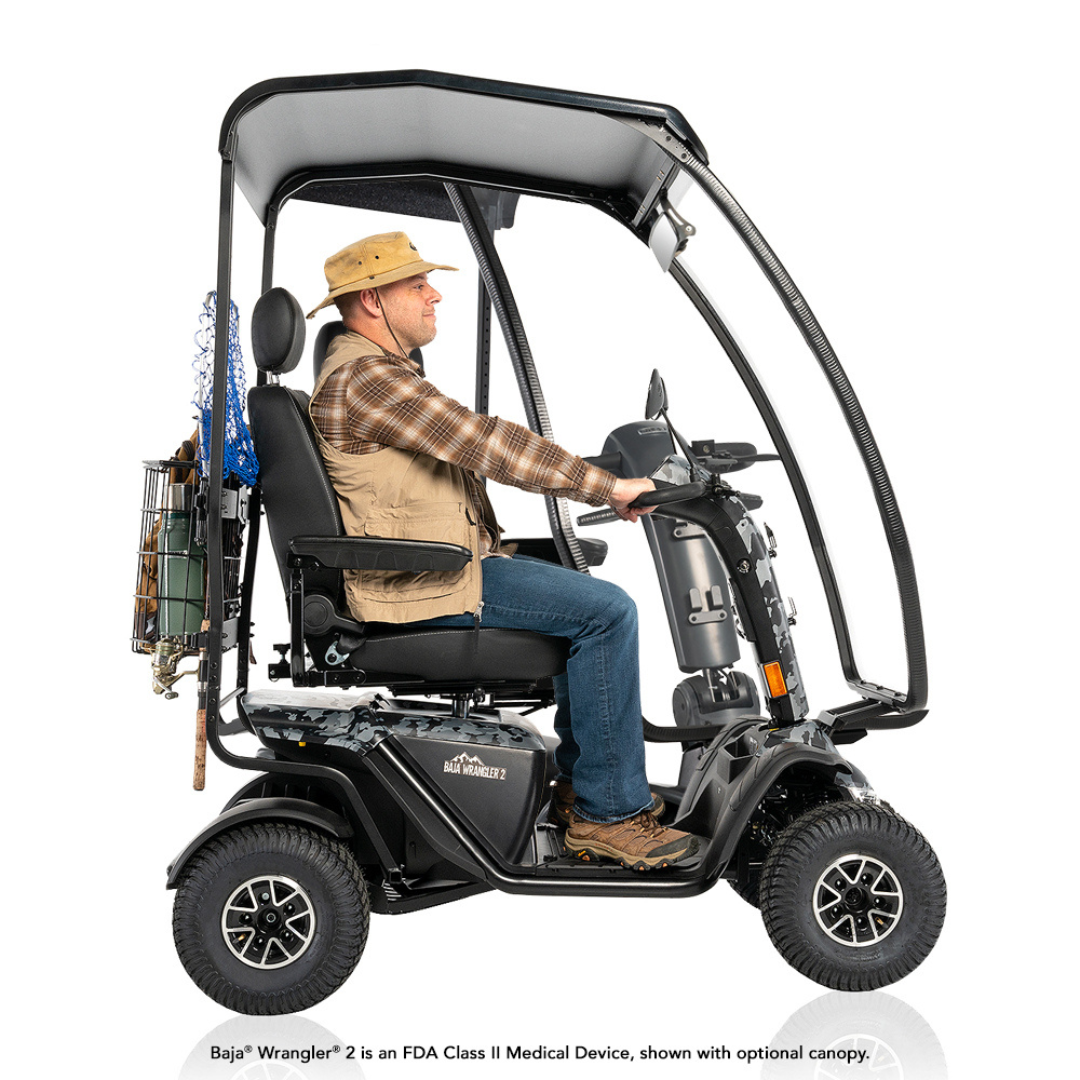 A man in outdoor gear sits on a Pride Baja® Wrangler® 2 Heavy Duty Outdoor Scooter with large wheels and a canopy. Fishing rods and a basket are attached to the back, highlighting its rugged performance for outdoor activities.
