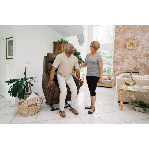 An older man stands up from a Pride Mobility VivaLift!® Tranquil 2 Recliner with Lift Assist, helped by a smiling woman in a bright, modern living room featuring large windows and stylish decor.