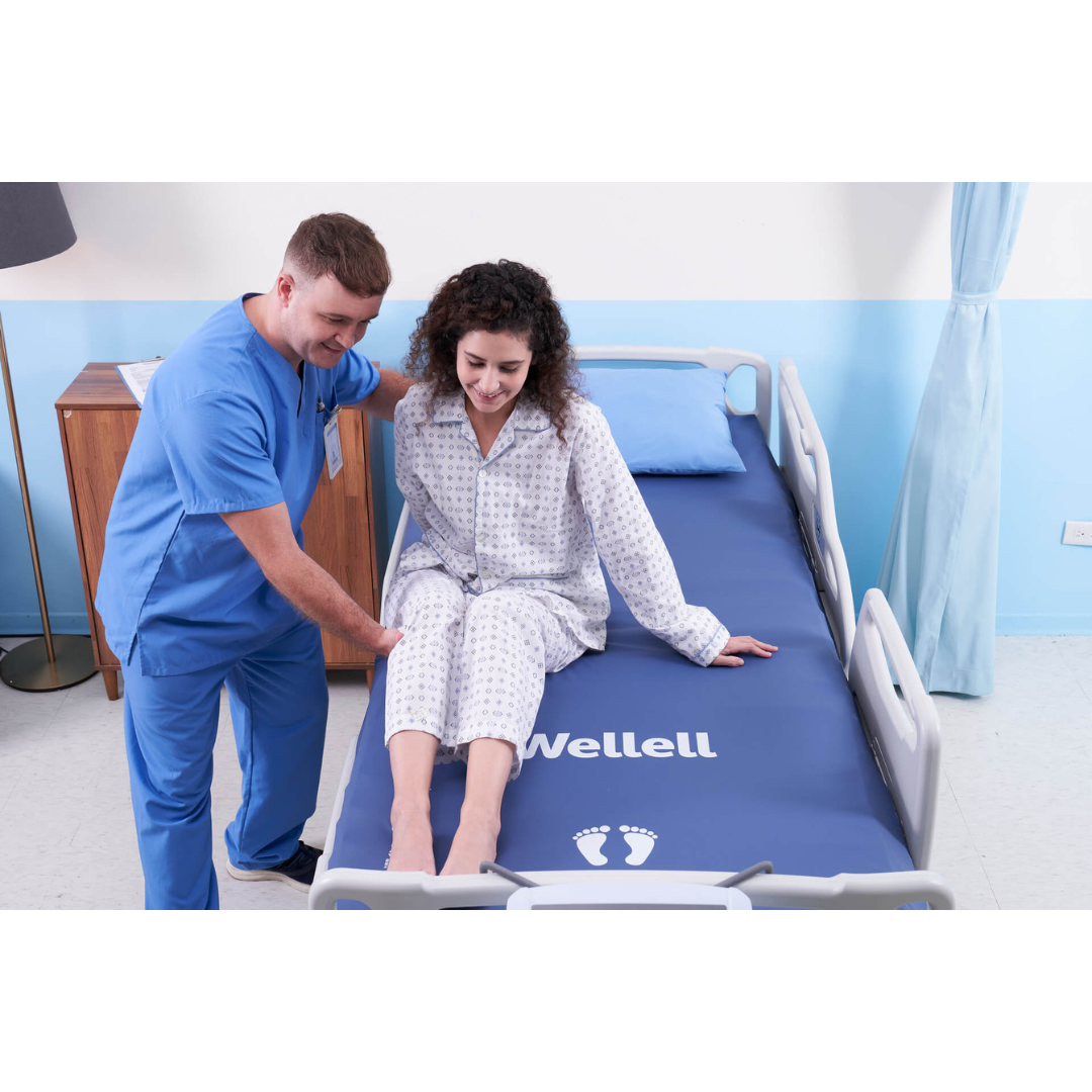 A nurse in blue scrubs assists a woman in pajamas as she sits on the edge of a hospital bed, equipped with the Wellell Procare Auto G2 | Alternating Pressure & LAL Mattress System, in a bright and clean hospital room.
