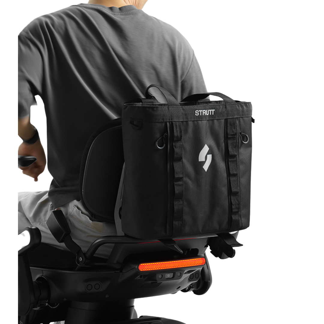 A person in a gray shirt rides a bike or scooter with the Rear Storage Bag | Strutt ev1 Compatible, featuring a logo and adjustable straps, attached behind the seat. The bag provides expanded space and convenient rear storage.