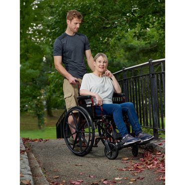 A man pushes a woman in a Drive Rebel Wheelchair with Quick-Release Wheels up a paved path bordered by trees. The woman smiles, and both enjoy the outdoors on a sunny day.