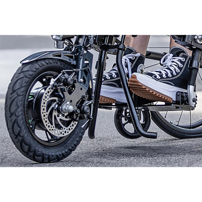 Close-up of a person wearing black and white sneakers with thick, wavy soles while riding a small black bicycle or using the Rehasense ECO Assist Power Chair Conversion System. The focus is on the shoes and front wheel.