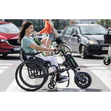 A woman in a manual wheelchair equipped with the Rehasense ECO Assist Power Chair Conversion System crosses a city street at a crosswalk, highlighting an innovative mobility solution among cars and cyclists.