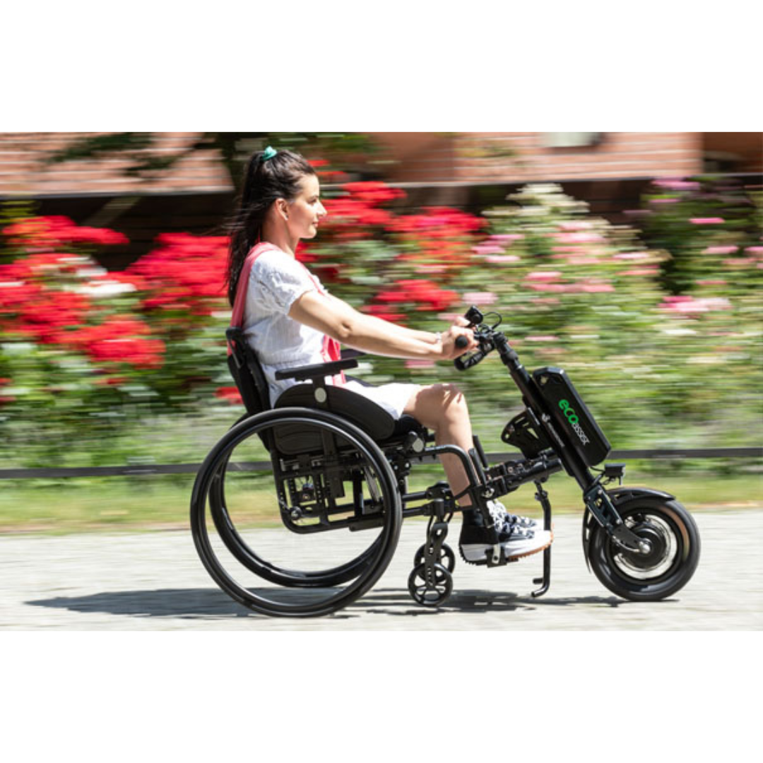 A woman rides outdoors in a manual wheelchair with the Rehasense ECO Assist Power Chair Conversion System, surrounded by blurred red and pink flowers, highlighting the added movement and speed this innovative mobility solution provides.