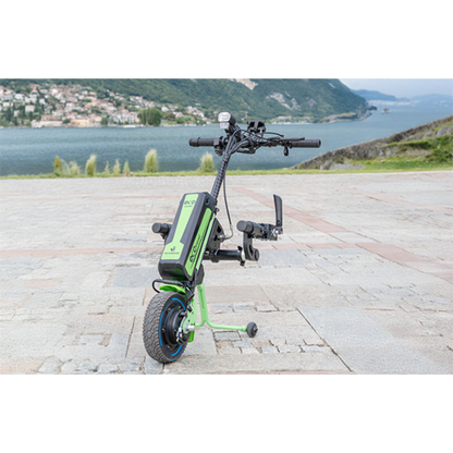 A Rehasense ECO Travel LX Power Add-On – Lightweight Wheelchair Motor Assist is attached to a manual wheelchair parked on paving near a large body of water, with hills and buildings visible in the background.