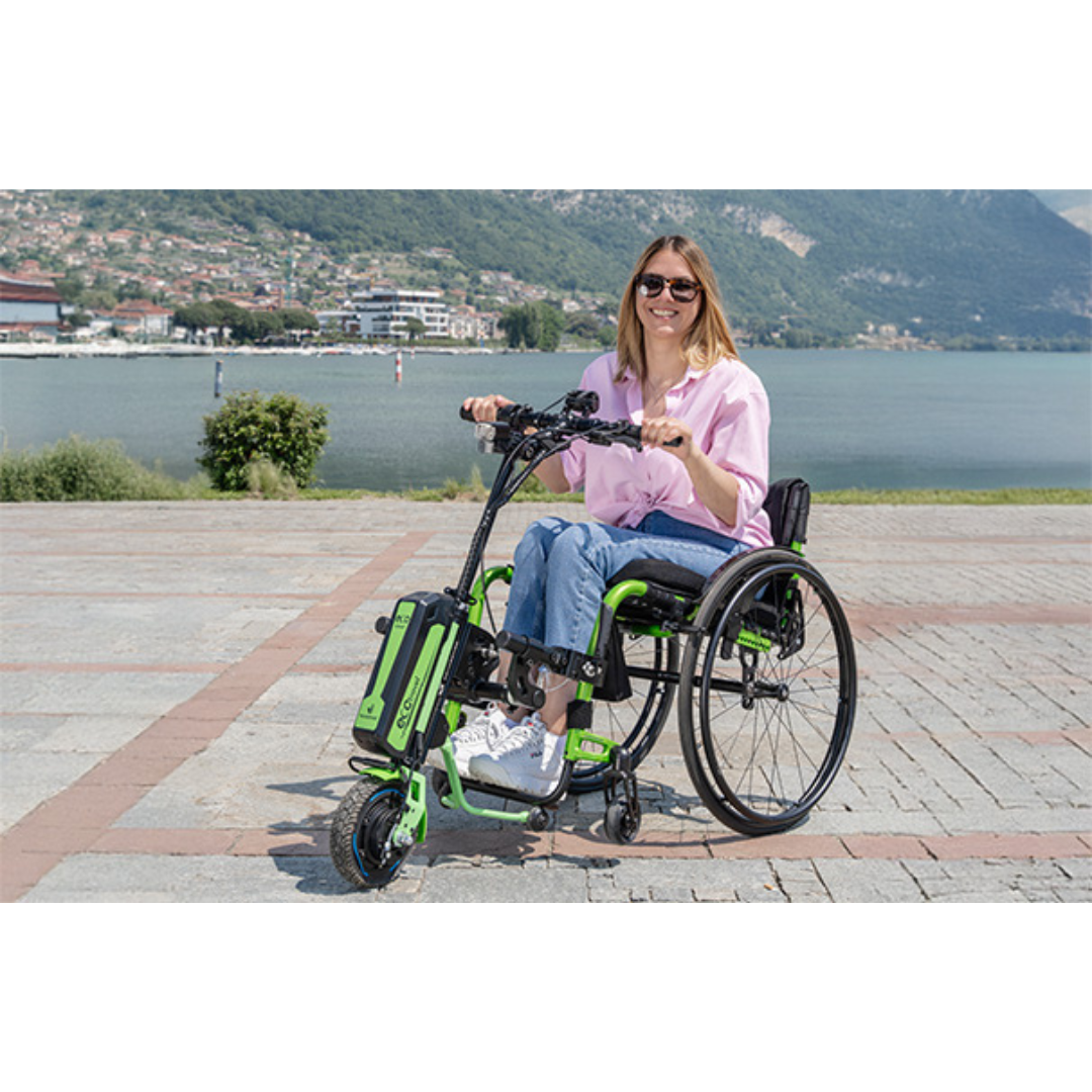 A woman in sunglasses and a pink shirt sits in a manual wheelchair equipped with the Rehasense ECO Travel LX Power Add-On – Lightweight Wheelchair Motor Assist, smiling outdoors near a lake with mountains and buildings in the background.