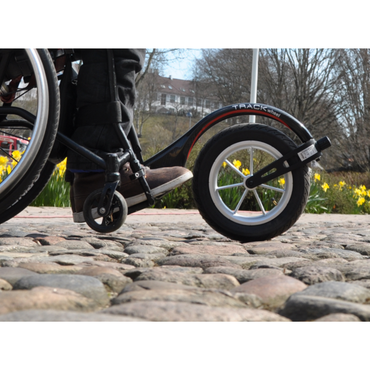 Close-up of a wheelchair user's feet on cobblestones with a Rehasense Single-Arm Track Wheel - XL 12" Carbon Outdoor Wheel attached. Yellow flowers, trees, and a distant building enhance the background scenery.
