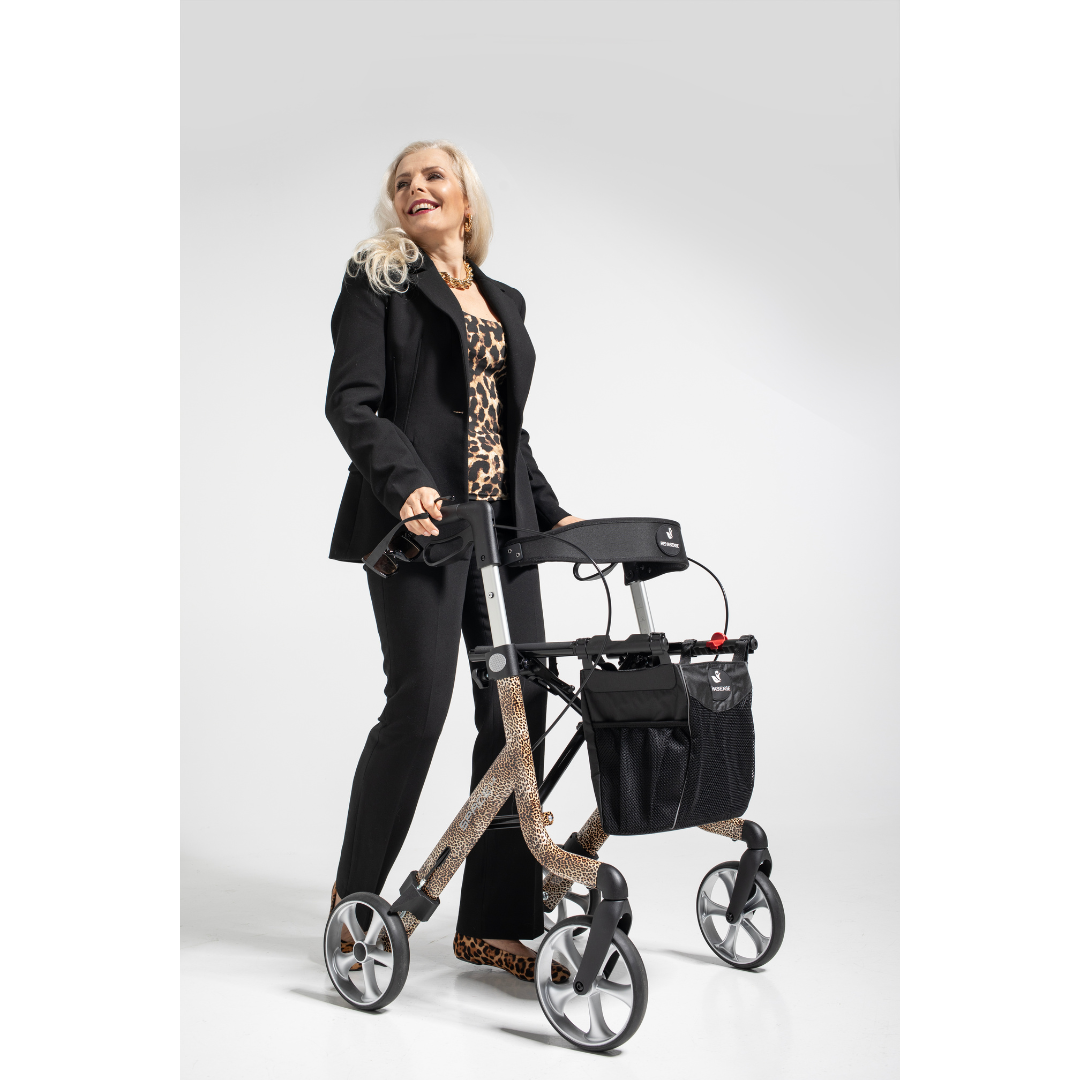 An older woman with long white hair smiles confidently while standing with a Rehasense Space LX Art Deco Ultralight Rollator featuring a leopard print, dressed in a black suit and matching leopard-print top and shoes against a plain white background.