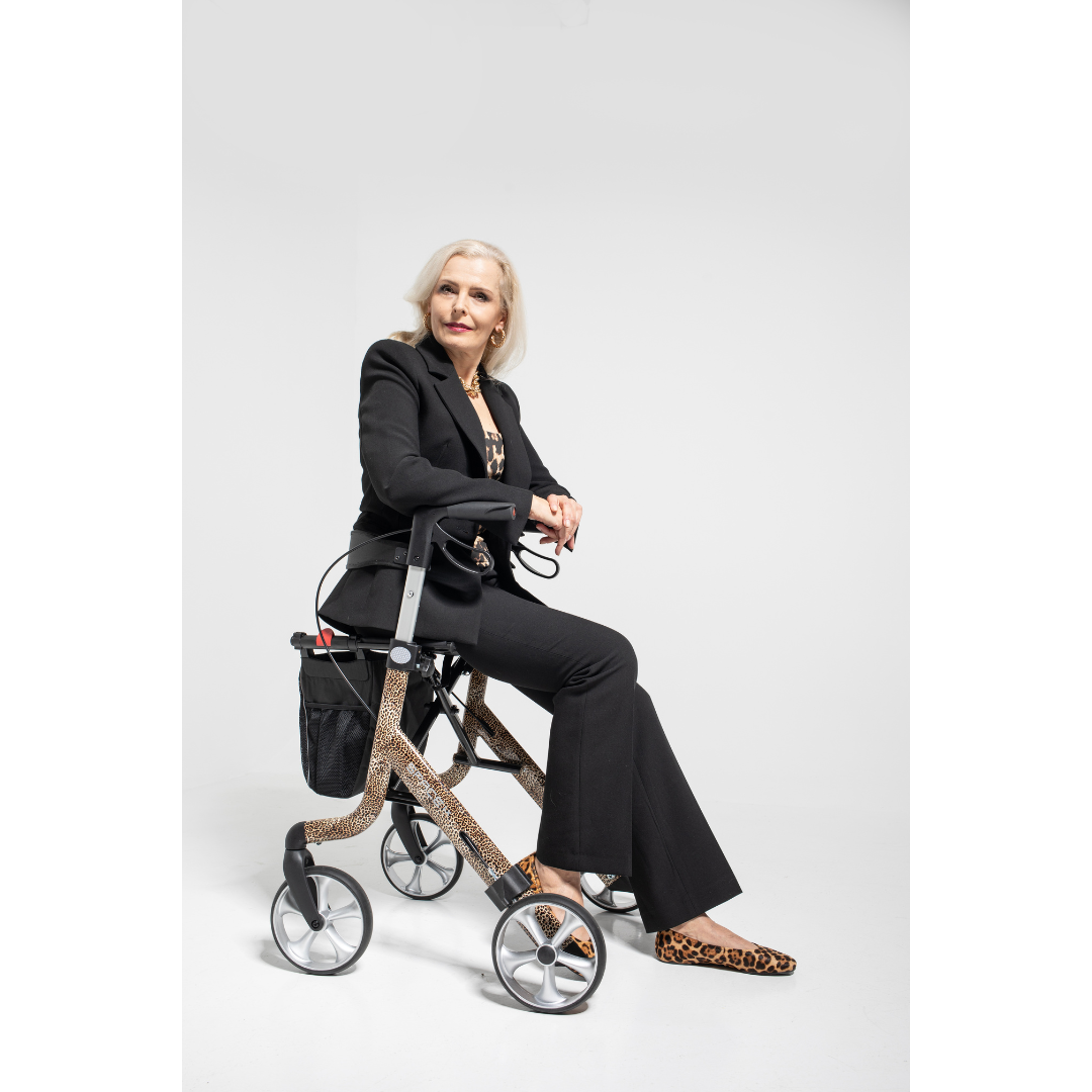 A stylish older woman with long blonde hair sits confidently on a Rehasense Space LX Art Deco Ultralight Rollator, dressed in a black suit and leopard-print shoes, against a plain white background.