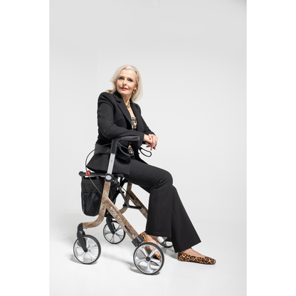 A stylish older woman with long blonde hair sits confidently on a Rehasense Space LX Art Deco Ultralight Rollator, dressed in a black suit and leopard-print shoes, against a plain white background.
