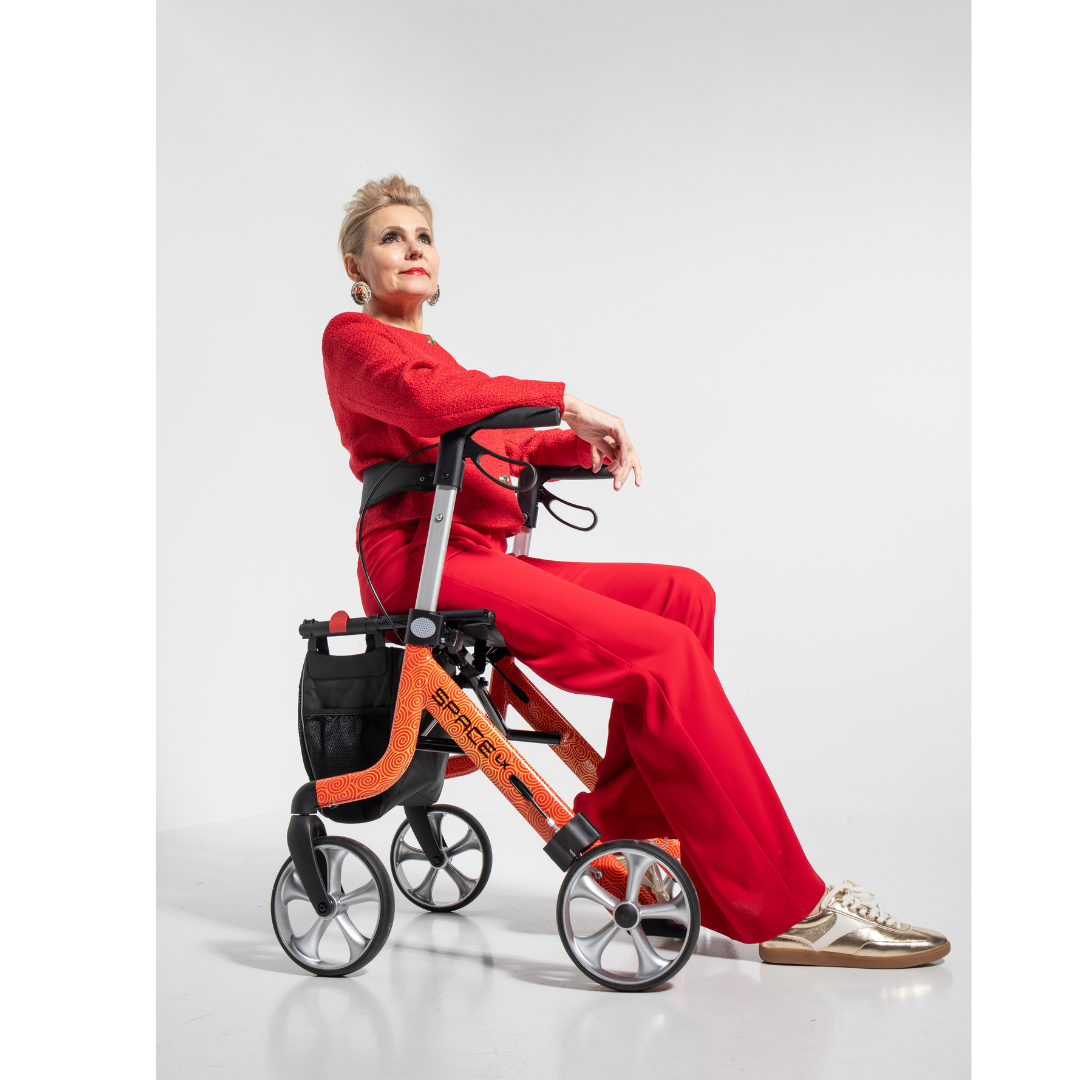 A stylish older woman in a red outfit sits confidently on a Rehasense Space LX Art Deco Ultralight Rollator with large wheels, gazing upward against a plain white background.