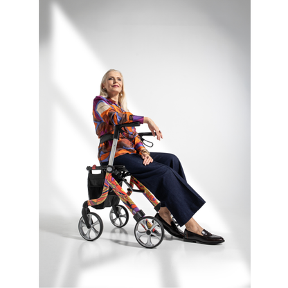An older woman with long blonde hair, in a colorful blouse and dark pants, sits confidently on a Rehasense Space LX Art Deco Ultralight Rollator in a bright studio, her smile reflecting the comfort of this lightweight aluminium rollator.