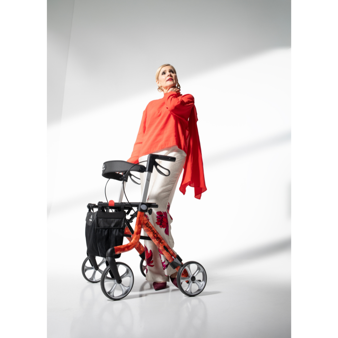 A stylish woman in a bright red top and floral pants stands confidently with the Rehasense Space LX Art Deco Ultralight Rollator in a brightly lit, minimalist setting.