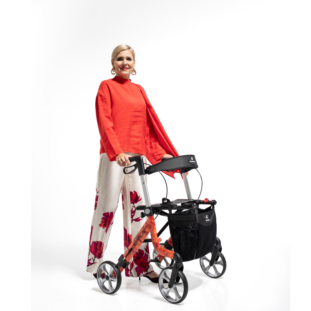 A smiling woman in a red long-sleeve top and white pants with red floral prints stands holding a Rehasense Space LX Art Deco Ultralight Rollator against a plain white background.