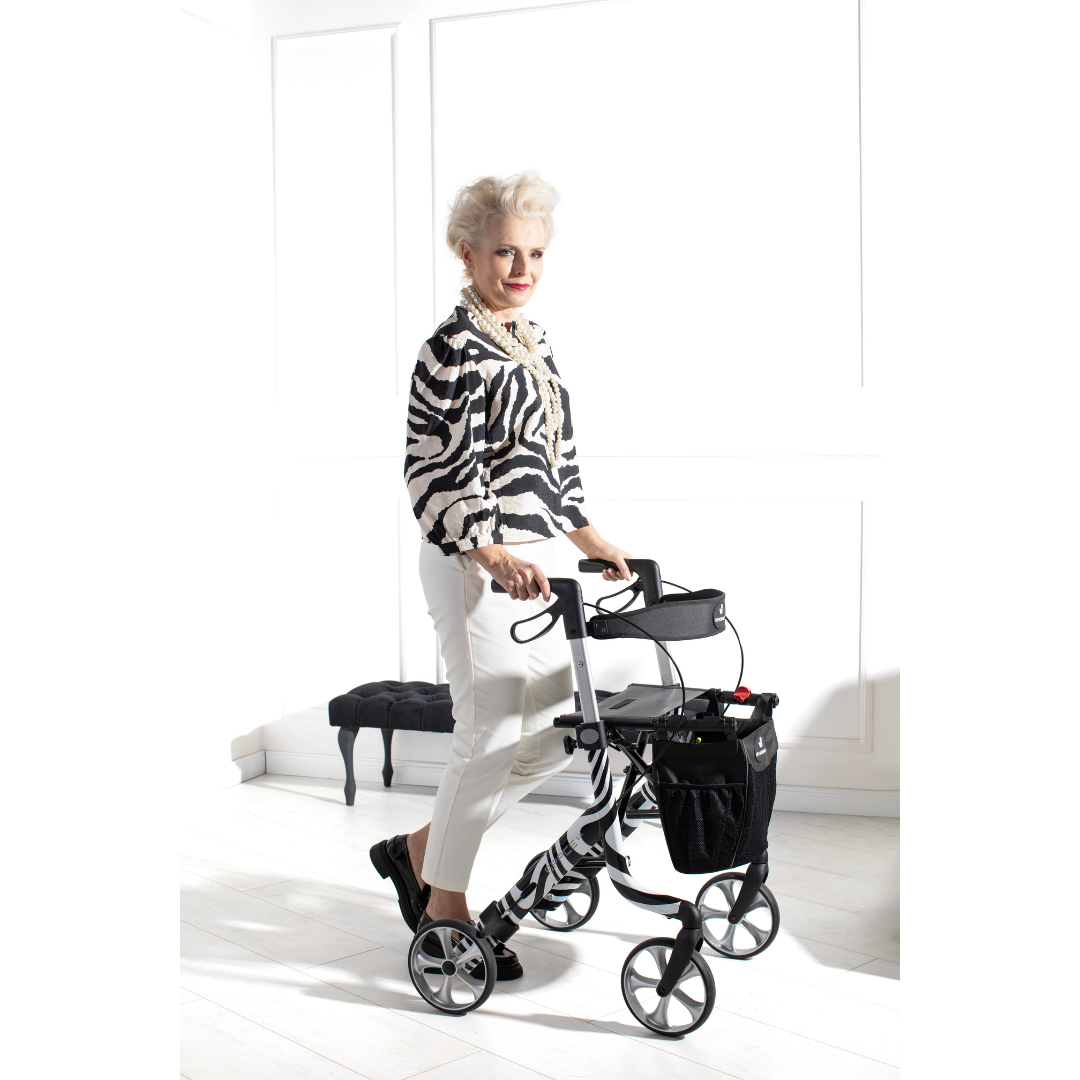 An older woman with white hair and a zebra-print top smiles indoors as she uses the Rehasense Space LX Art Deco Ultralight Rollator in a bright room with white walls and a small bench in the background.