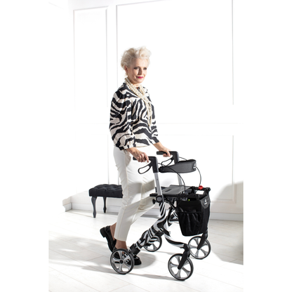 An older woman with white hair and a zebra-print top smiles indoors as she uses the Rehasense Space LX Art Deco Ultralight Rollator in a bright room with white walls and a small bench in the background.