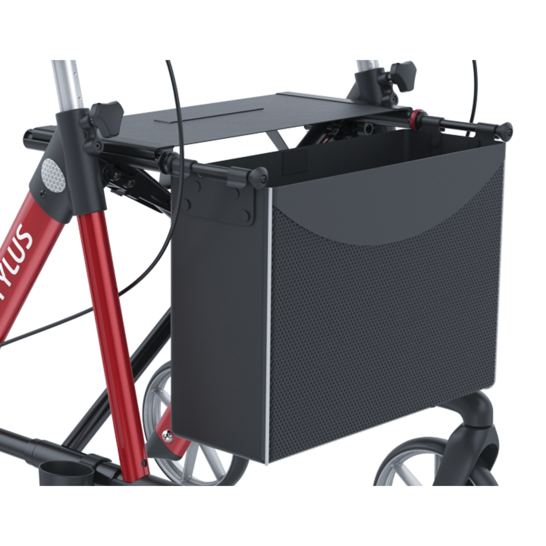 A close-up of a black storage basket attached to the front frame of a red and black Rehasense Stylus Premium Indoor & Outdoor Rollator with three visible wheels.