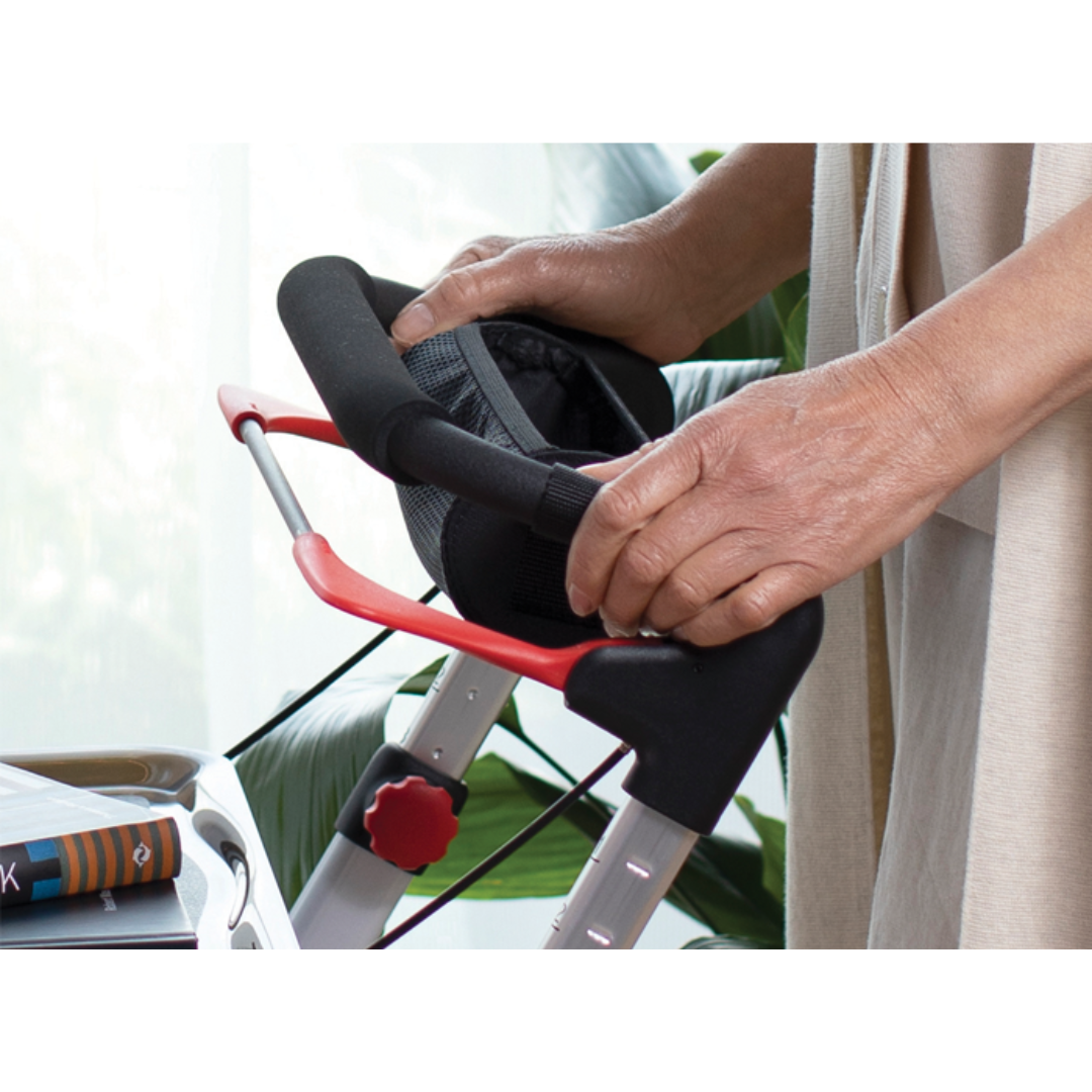 A close-up of an older adult’s hands gripping the handles of a Rehasense Pixel Indoor Rolling Walker, with a tray and books nearby. The indoor scene includes some greenery in the background.