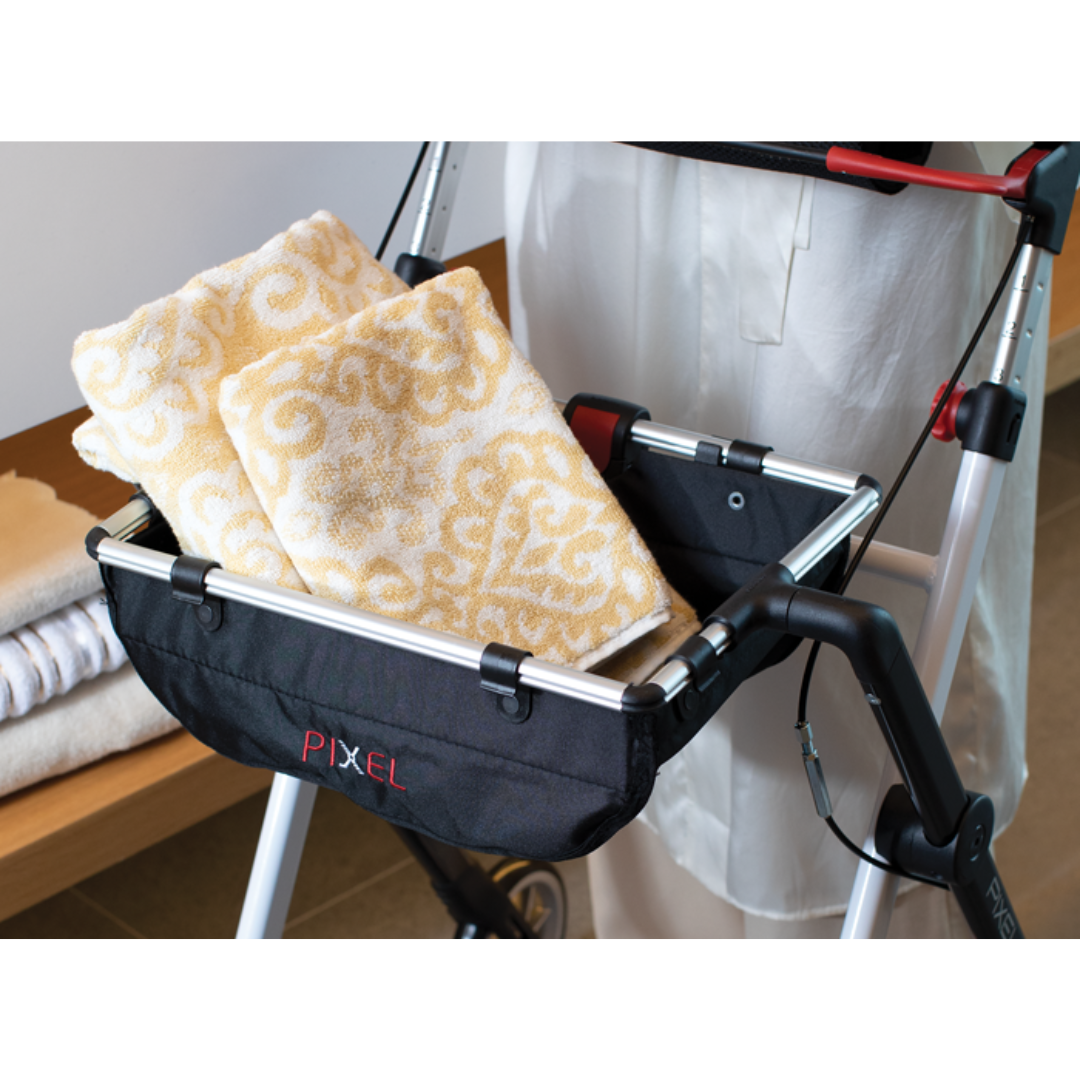 A person stands behind a black Rehasense Pixel Indoor Rolling Walker with a basket containing two folded yellow and white towels. Additional folded towels are stacked on a nearby bench.