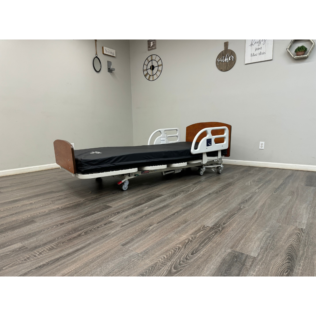 A Med-Mizer RetractaBed Bariatric Hi/Low Electric Bed with width adjustment, wooden head and footboards, safety rails, and black mattress stands in an empty room with light gray walls, wood-patterned floor, wall art, and plants.