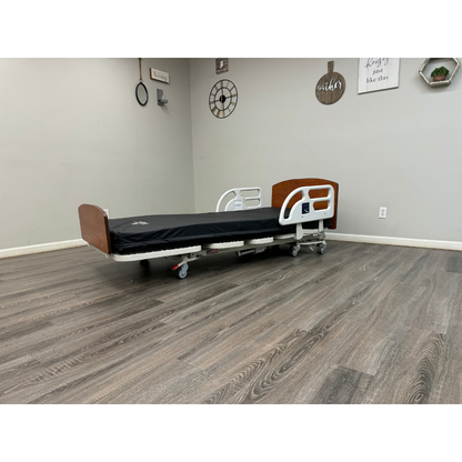 A Med-Mizer RetractaBed Bariatric Hi/Low Electric Bed with width adjustment, wooden head and footboards, safety rails, and black mattress stands in an empty room with light gray walls, wood-patterned floor, wall art, and plants.