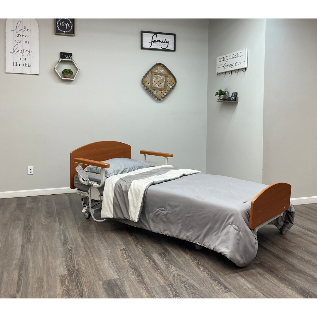 The Med-Mizer RetractaBed Bariatric Hi/Low Electric Bed, with gray bedding, features width adjustment and safety rails. It sits in a clean room with wood floors, light gray walls, wall art, shelves with plants, and positive quote decor.