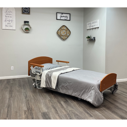 The Med-Mizer RetractaBed Bariatric Hi/Low Electric Bed, with gray bedding, features width adjustment and safety rails. It sits in a clean room with wood floors, light gray walls, wall art, shelves with plants, and positive quote decor.
