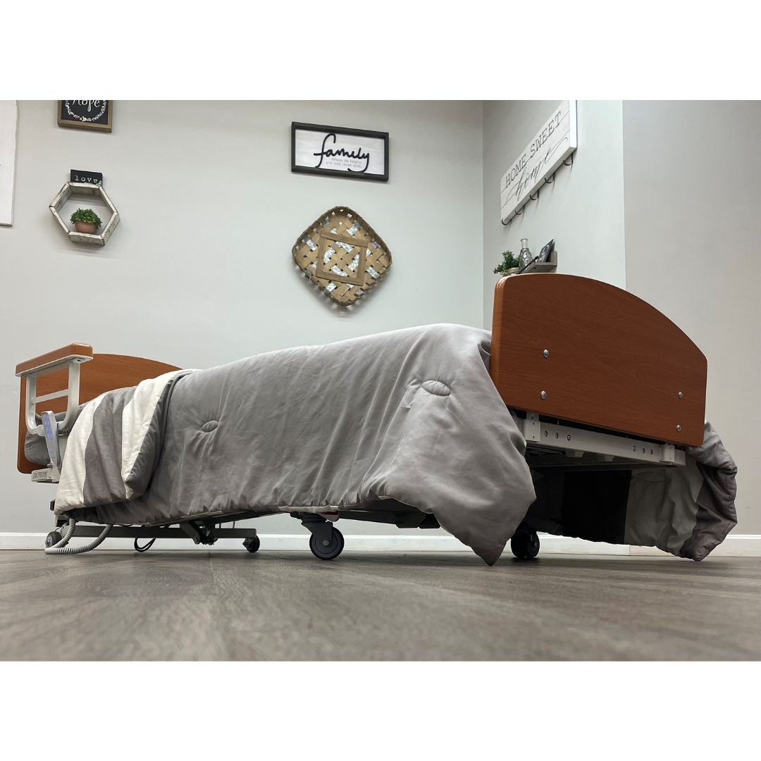 A low-angle view of the Med-Mizer RetractaBed Bariatric Hi/Low Electric Bed with safety rails, gray blanket, wood headboards, and wheels in a decorated room with wall art and wooden floors; bed width is adjustable for space-saving.