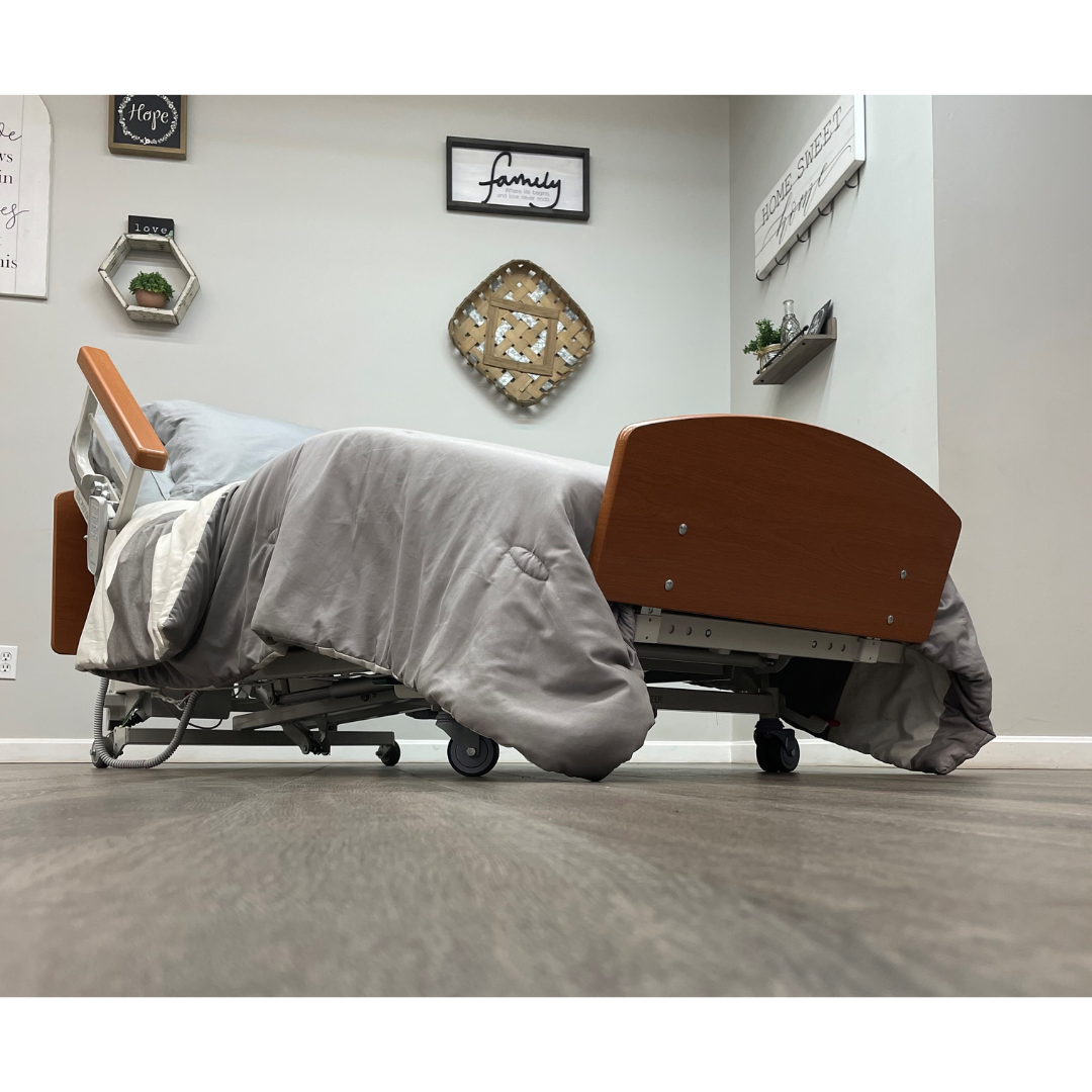 A low-angle view of the Med-Mizer RetractaBed Bariatric Hi/Low Electric Bed with width adjustment, featuring gray bedding in a stylish room. The wood floor matches the bed’s wheels, wooden headboard and footboard, and safety rails.