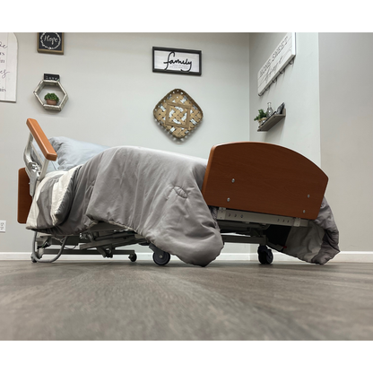 A low-angle view of the Med-Mizer RetractaBed Bariatric Hi/Low Electric Bed with width adjustment, featuring gray bedding in a stylish room. The wood floor matches the bed’s wheels, wooden headboard and footboard, and safety rails.