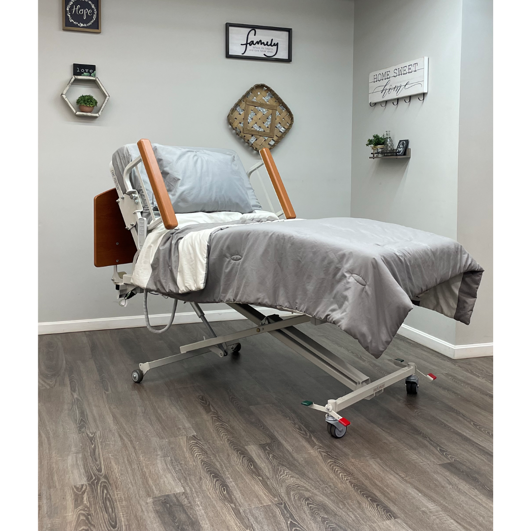 The Med-Mizer RetractaBed Bariatric Hi/Low Electric Bed, with width adjustment and safety rails, is set in a modern room with gray bedding, wooden floors, wall decor, and shelves holding plants. The head of the bed is partially elevated.