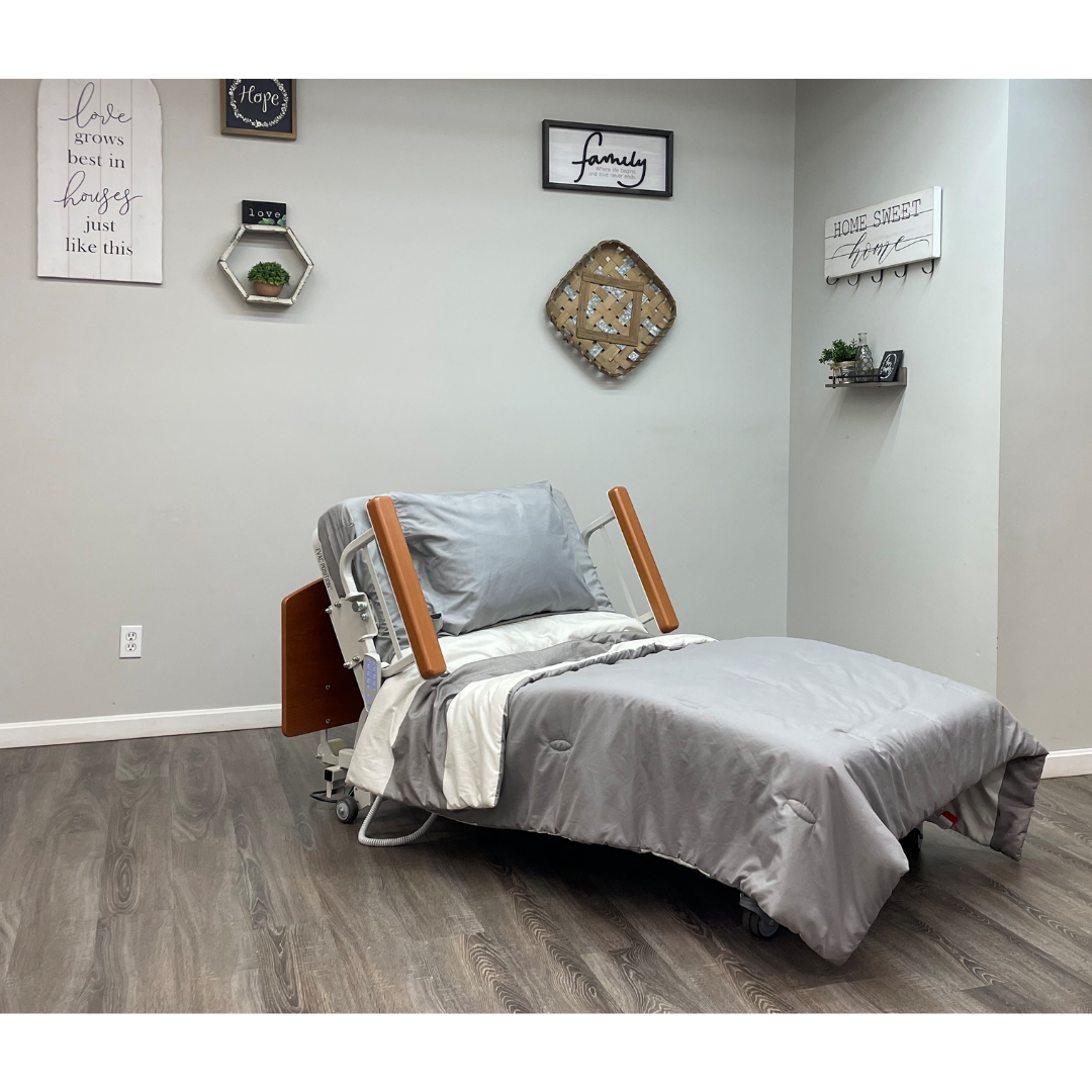A Med-Mizer RetractaBed - Bariatric Hi/Low Electric Bed - Width Adjustable with gray bedding sits in a cozy room with wooden floors and wall art, blending space-saving hospital bed design and homelike comfort.