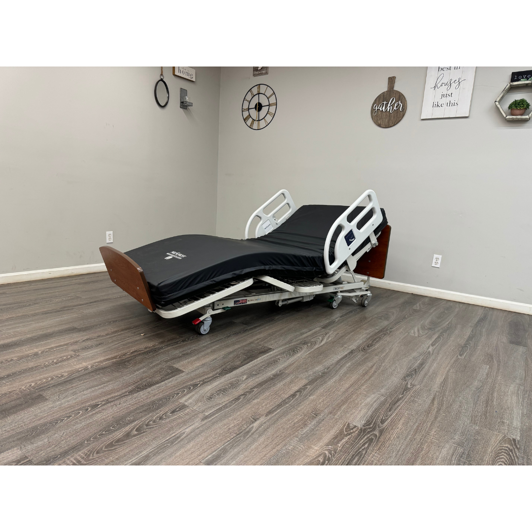 A Med-Mizer RetractaBed Bariatric Hi/Low Electric Bed with black mattress, white safety rails, and wood head/footboards is in a room with light gray walls, wood flooring, and decorative wall art.