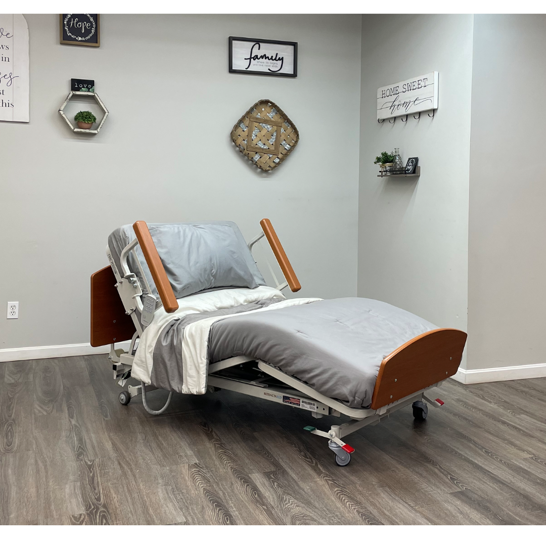 A Med-Mizer RetractaBed Bariatric Hi/Low Electric Bed (width adjustable) with wooden ends and gray bedding sits in a cozy room with light walls, wood floors, wall art, shelves, and built-in safety rails for comfort and security.