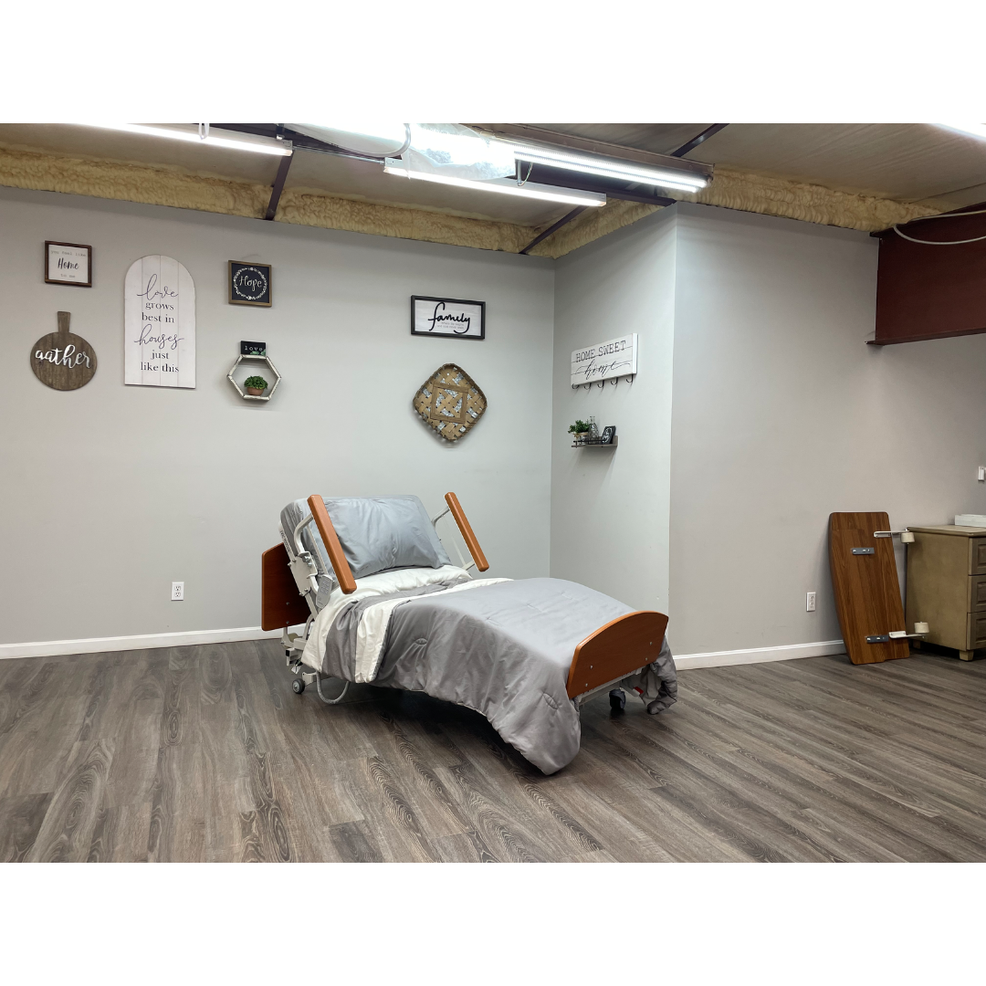 A Med-Mizer RetractaBed Bariatric Hi/Low Electric Bed with width adjustment and sturdy safety rails sits in a clean room with gray bedding, wood floors, neutral walls, inspirational wall art, and a plant shelf.