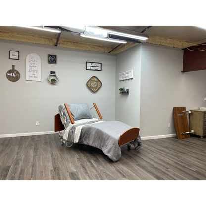 A Med-Mizer RetractaBed Bariatric Hi/Low Electric Bed with width adjustment and sturdy safety rails sits in a clean room with gray bedding, wood floors, neutral walls, inspirational wall art, and a plant shelf.