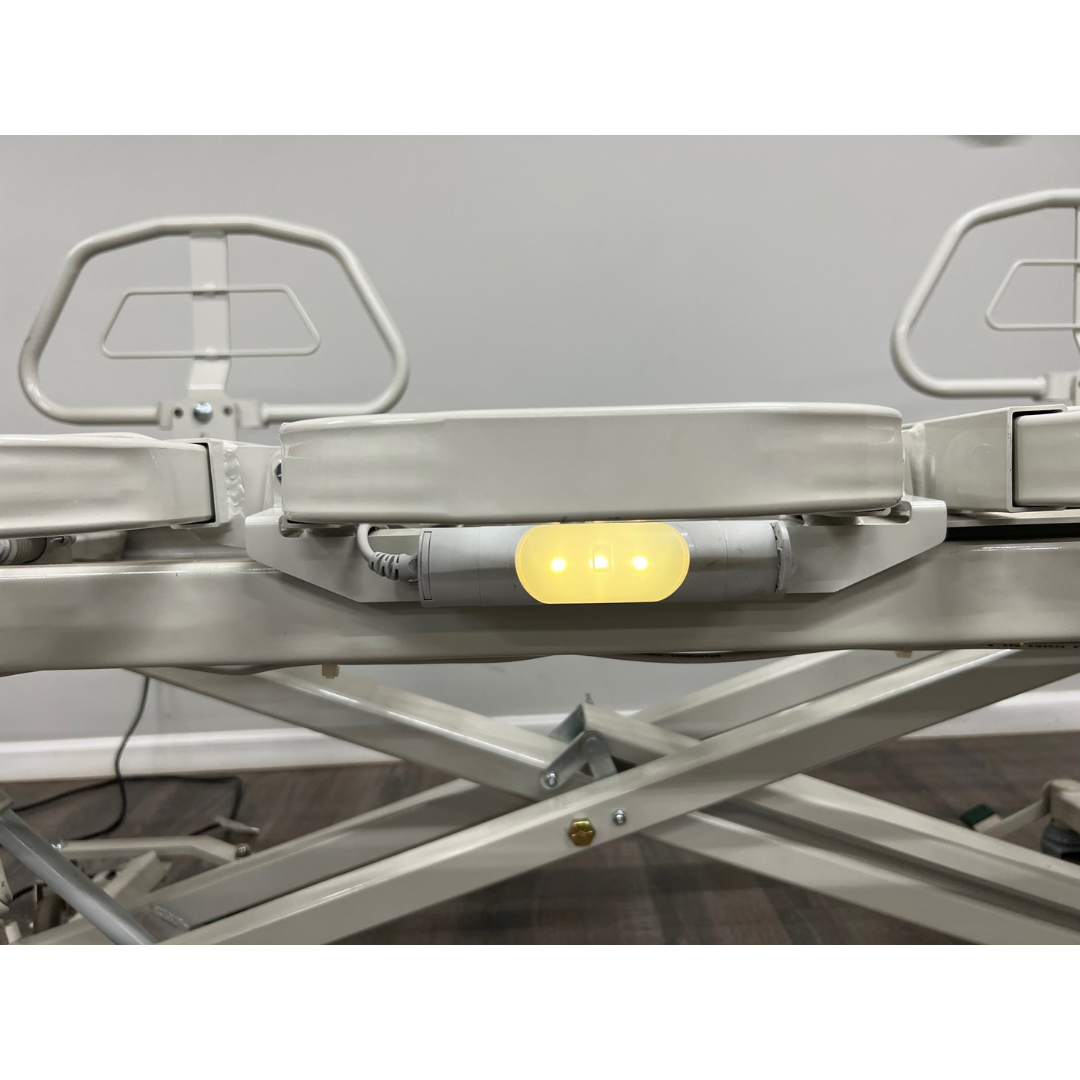 A close-up of the Med-Mizer RetractaBed - Bariatric Hi/Low Electric Bed - Width Adjustable, featuring an illuminated yellow indicator light, metal frame, and safety rails against a plain gray wall.