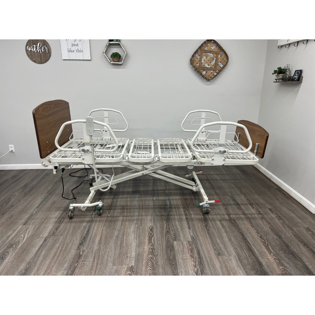The Med-Mizer RetractaBed Bariatric Hi/Low Electric Bed, with safety rails and wood accents, stands elevated and bedding-free in a room with gray walls, wood flooring, and decorative wall shelves for a clean, space-saving appearance.