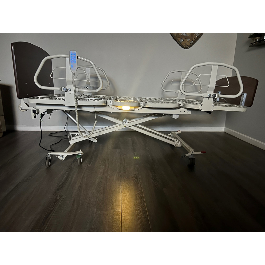 A Med-Mizer RetractaBed Bariatric Hi/Low Electric Bed with adjustable width, raised safety rails, an attached remote, and an under-bed light sits on a dark wood floor in a room with neutral walls.