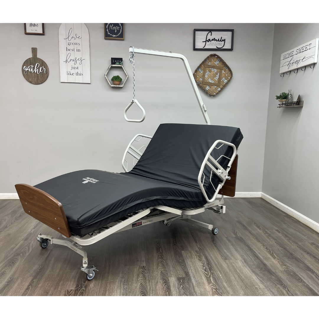 The Med-Mizer RetractaBed Bariatric Hi/Low Electric Bed with adjustable width, head and leg sections, safety rails, dark mattress, wooden head/footboards, and overhead trapeze bar is set in a room with wood flooring and decorative wall signs.