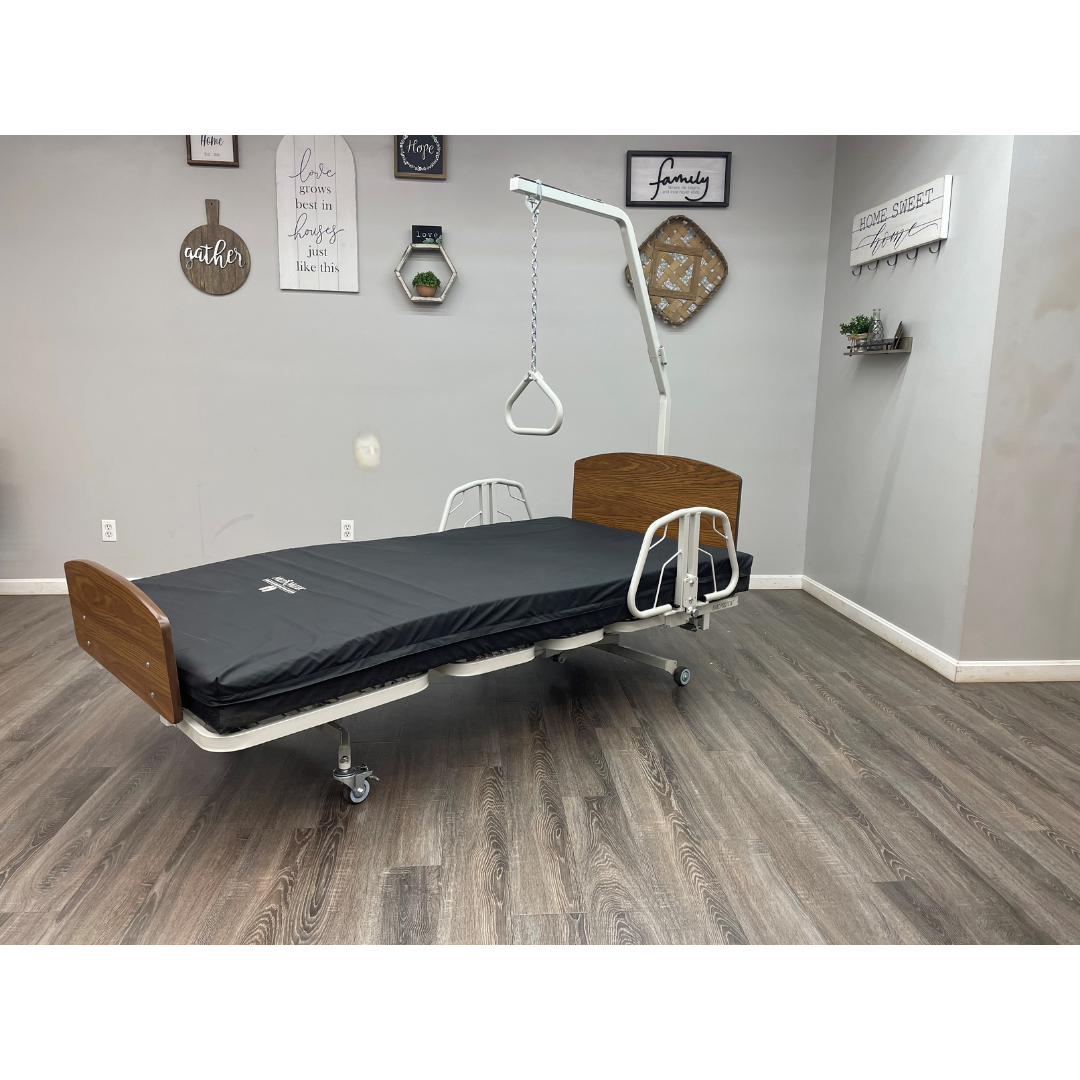 A Med-Mizer RetractaBed – Bariatric Hi/Low Electric Bed (Width Adjustable) with wooden headboard, footboard, safety rails, black mattress, and overhead trapeze bar is set in a gray-walled room with wood flooring and wall art.