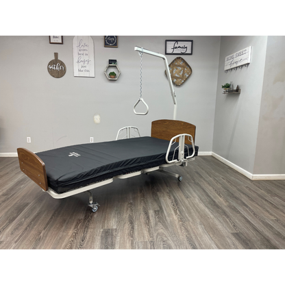 A Med-Mizer RetractaBed – Bariatric Hi/Low Electric Bed (Width Adjustable) with wooden headboard, footboard, safety rails, black mattress, and overhead trapeze bar is set in a gray-walled room with wood flooring and wall art.