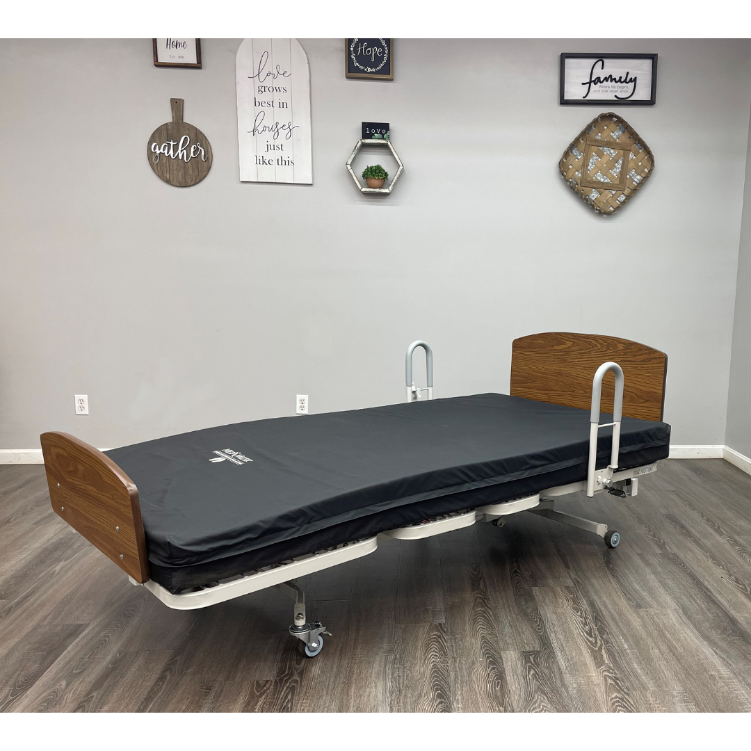 The Med-Mizer RetractaBed Bariatric Hi/Low Electric Bed with width adjustment, safety rails, black mattress, and wooden headboard/footboard stands on wheels in a room with wood flooring and decorative wall art.