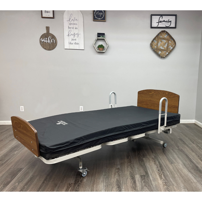 The Med-Mizer RetractaBed Bariatric Hi/Low Electric Bed with width adjustment, safety rails, black mattress, and wooden headboard/footboard stands on wheels in a room with wood flooring and decorative wall art.